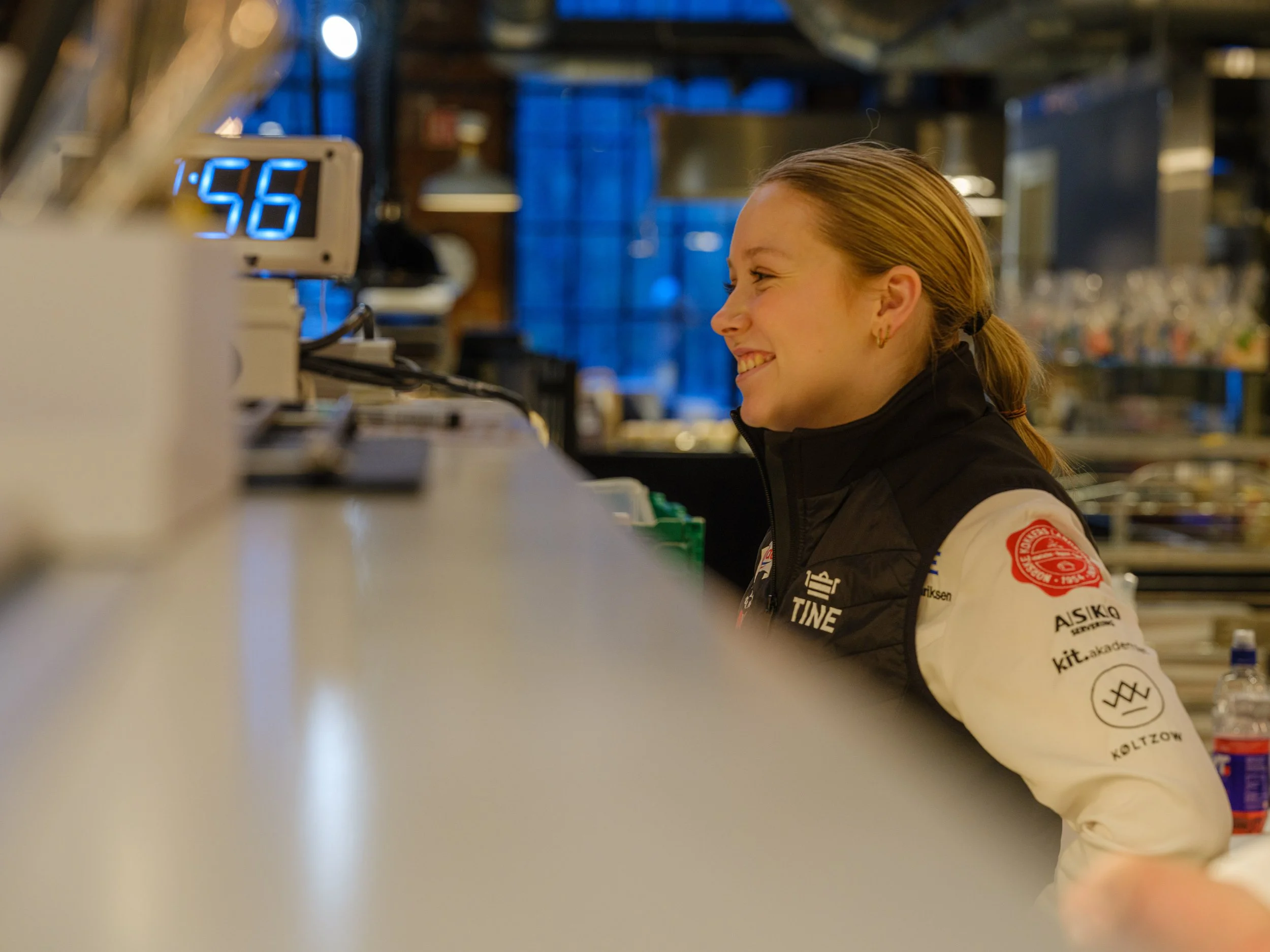 En kvinne smiler mens hun jobber på en bar eller kafé, med et digitalt tellerskilt som viser 1:55 i bakgrunnen.