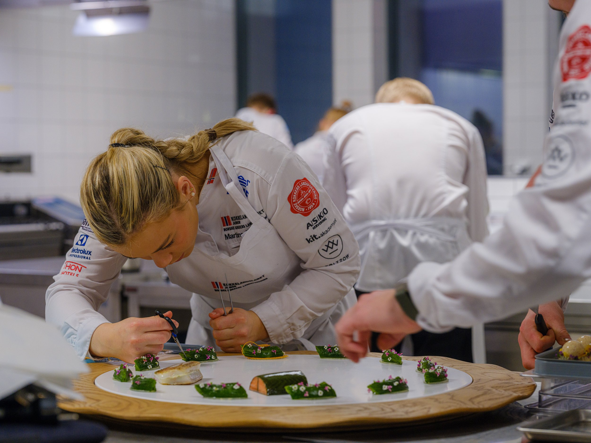 Kjøkkensjeferarbeid i et profesjonelt kjøkken, der de dekorerer en stor tallerken med små matretter, alle iført hvite kokkejakker.