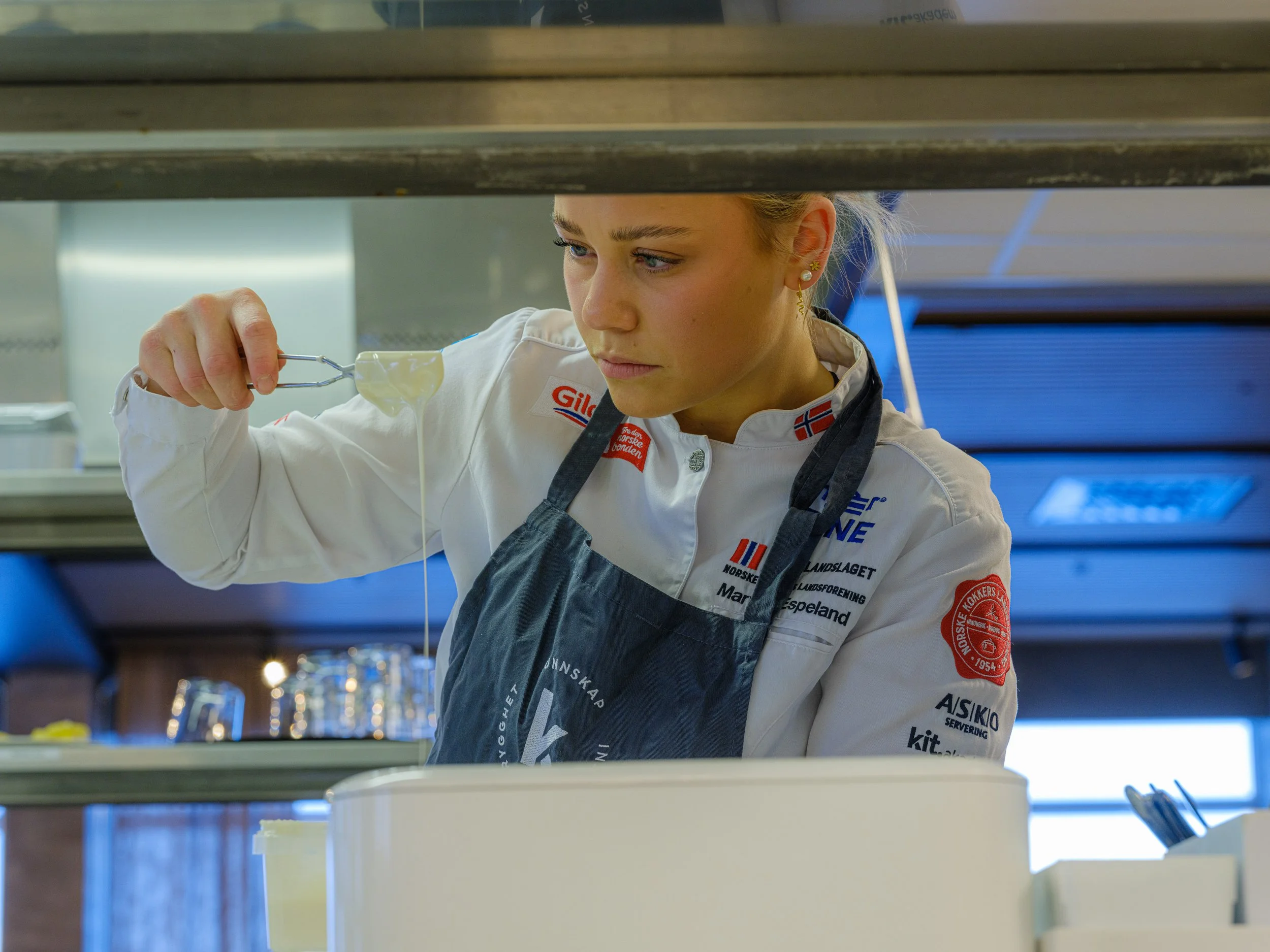 En kvinne som lager mat i et kjøkken, holder en skje med ost. Hun har på seg en hvit kokkejakke med norske flagg og sponsorlogoer, og en mørkeblå forkle.