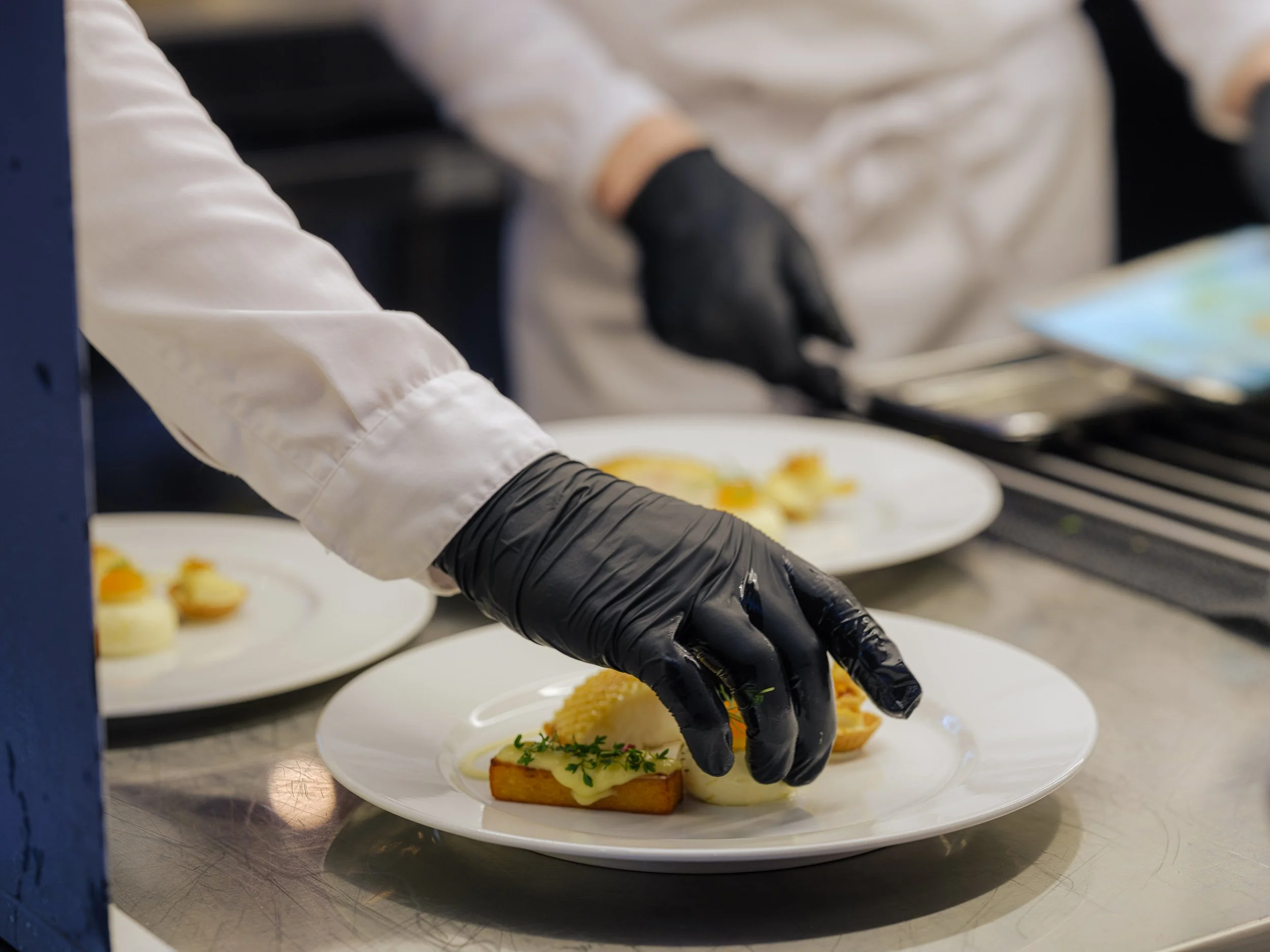 En kokk er i ferd med å legge opp et ferdig anrettet måltid på en hvit tallerken, med en brødskive med smør og urter, ved en kjøkkenbenk. Kokken bruker svarte hansker og en hvit kjøkkenhage.