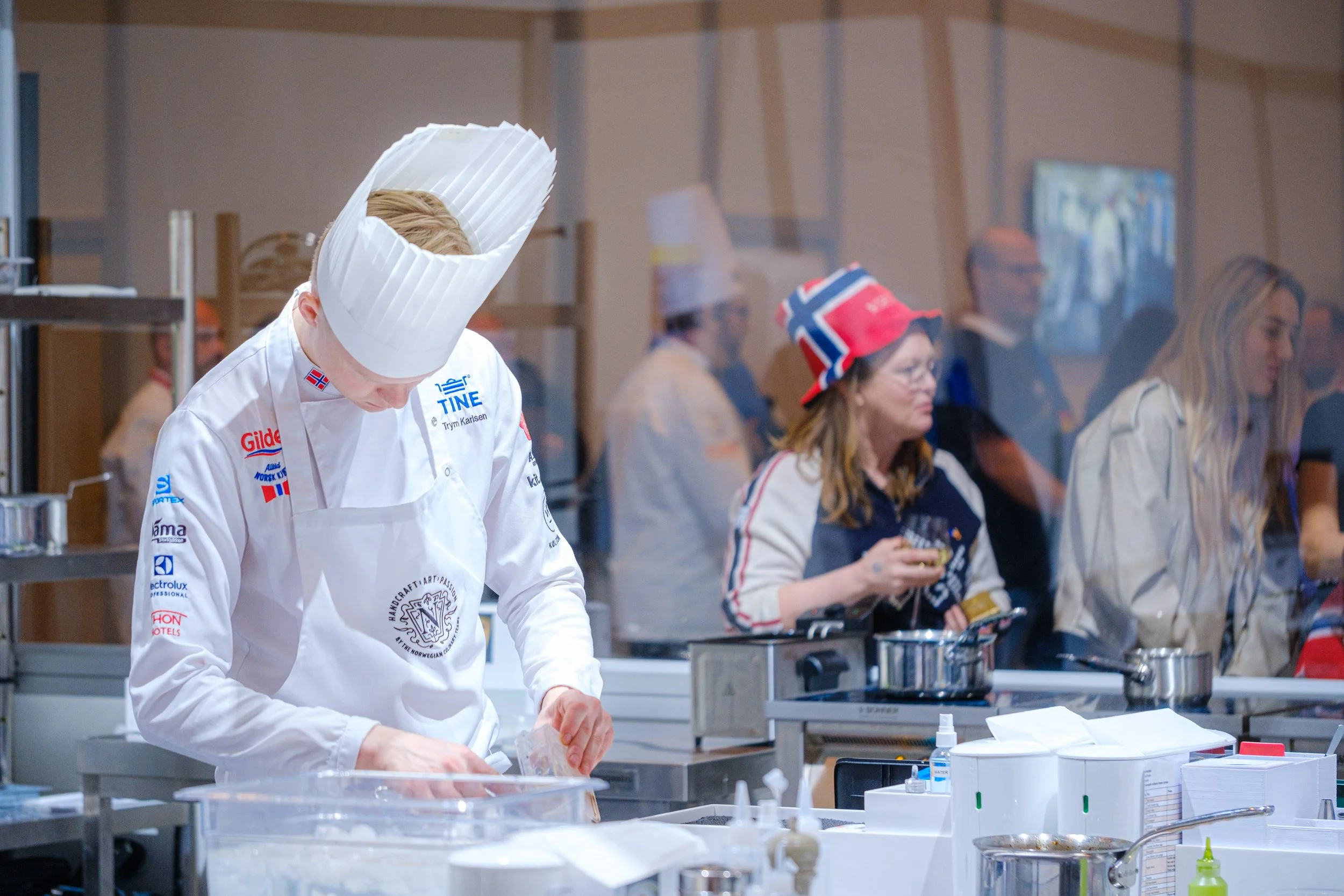 En kok i hvit uniform og kokkehatt jobber på kjøkkenet, mens folk bak glasset ser på, noen med norske flagg og cap, og andre som ser ut til å ha på seg kokkehatt og uniform.
