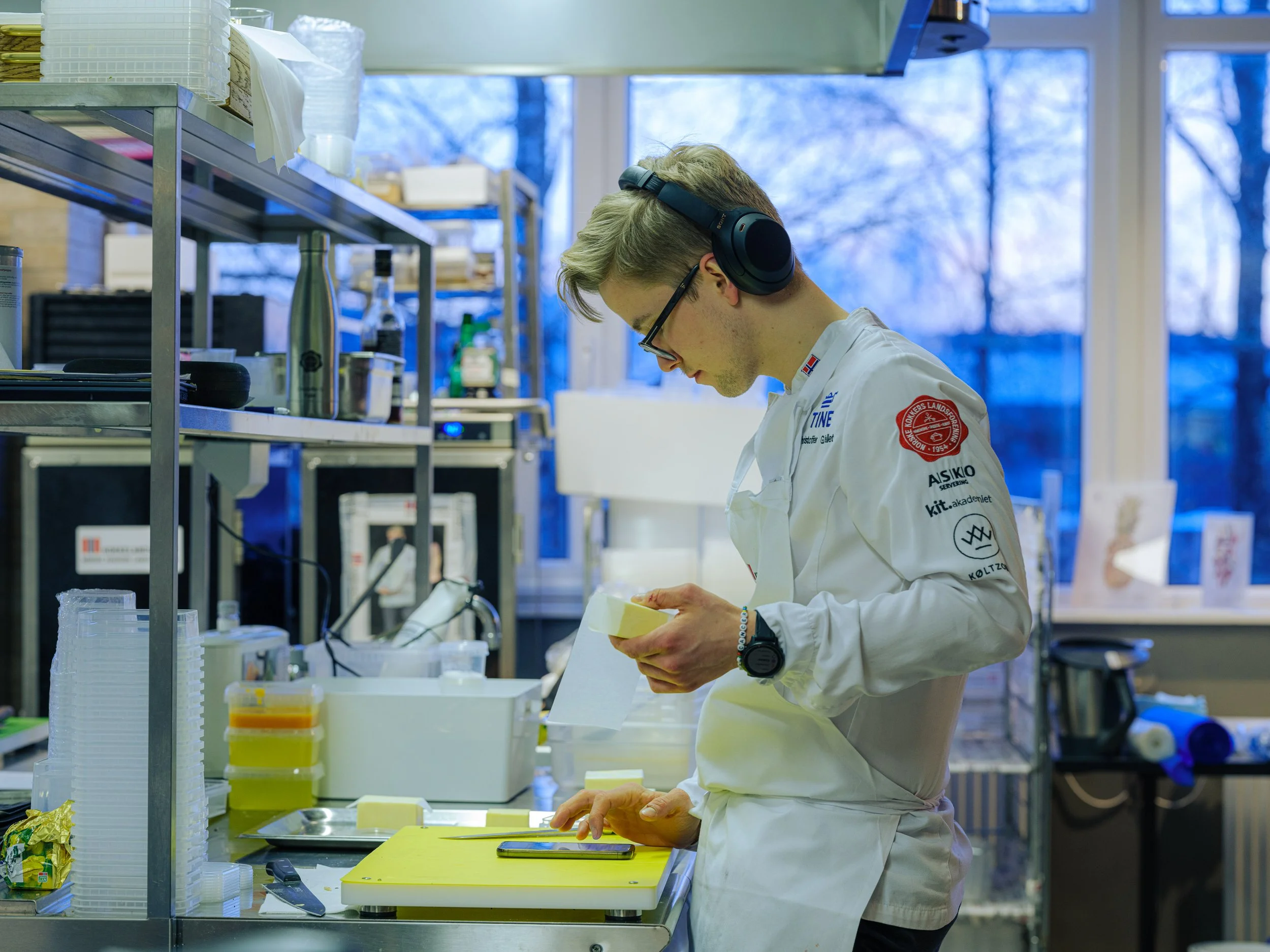 En ung mann i hvit kokkekjole med logo og patcher, med påsatt headset, står på et kjøkken og skjærer i en gul skjærefjøl. Han er innstilt og fokuserer på arbeidet sitt med en kniv. Det er masse kjøkkenutstyr og ulike beholdere rundt ham, og store vinduer i bakgrunnen viser snø og trær.