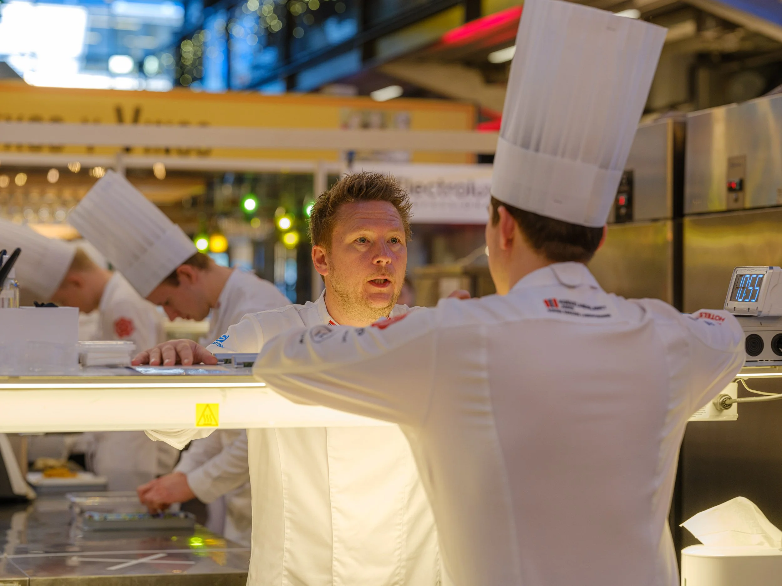 To kokker i hvite kokkeklær og høye kokkehatter snakker med hverandre i en restaurantkjøkkenet.