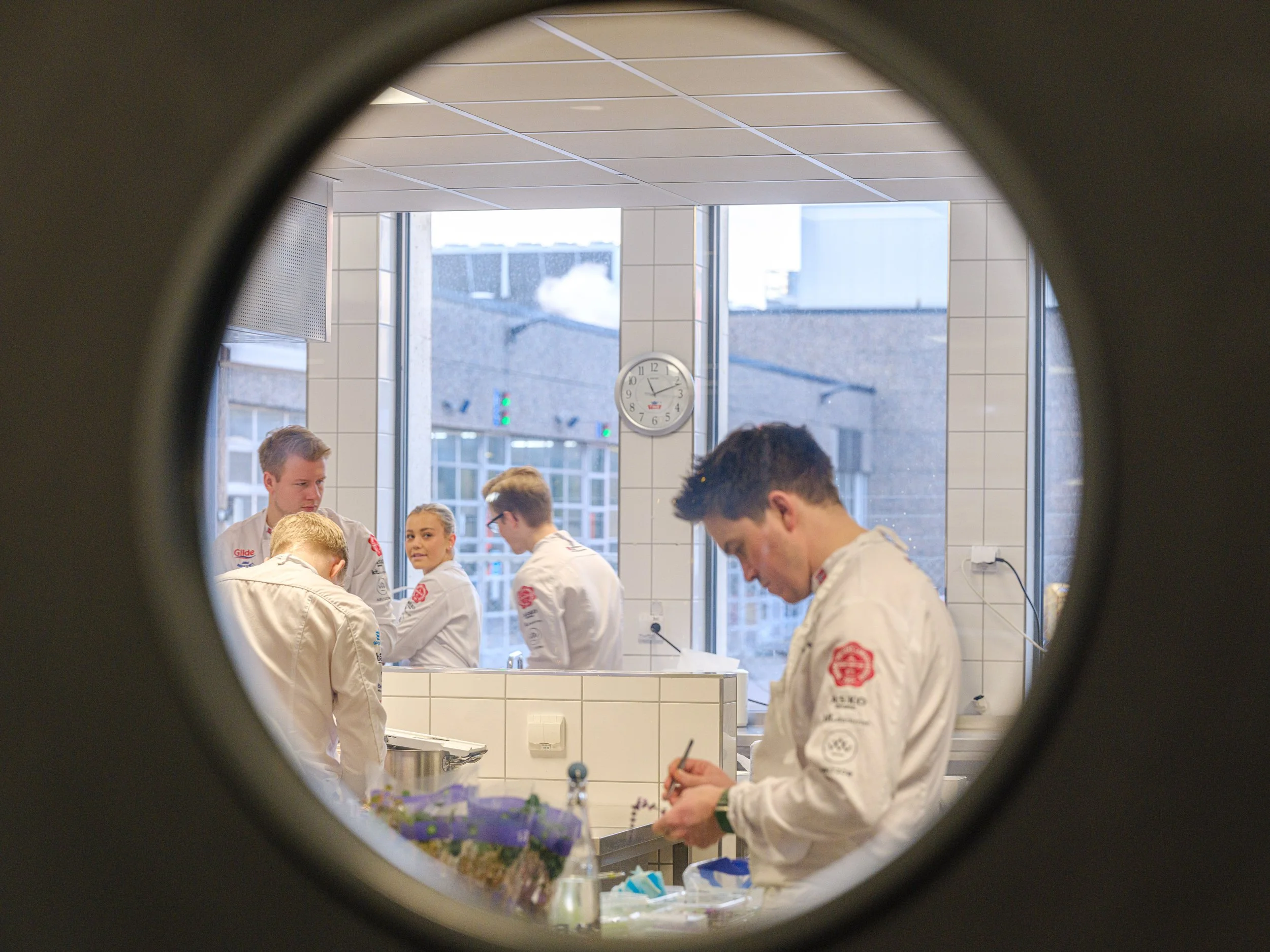 Kjøkkenmedarbeidere i kokkehatt og uniform som jobber på kjøkken gjennom et vindu.