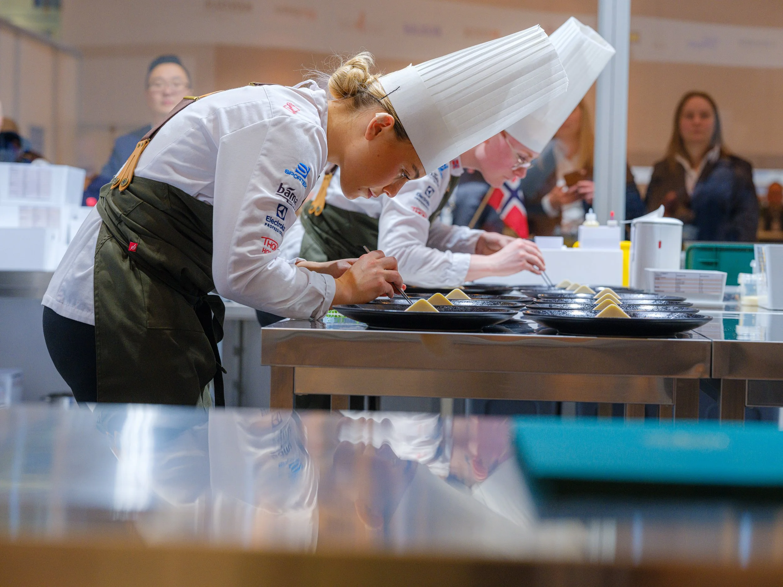 Kokkekonkurranse hvor to kokker med hvite kockhatter og hvide uniformer legger anretninger på tallerkener ved en arbeidsbenk. Noen tilskuere observerer i bakgrunnen.