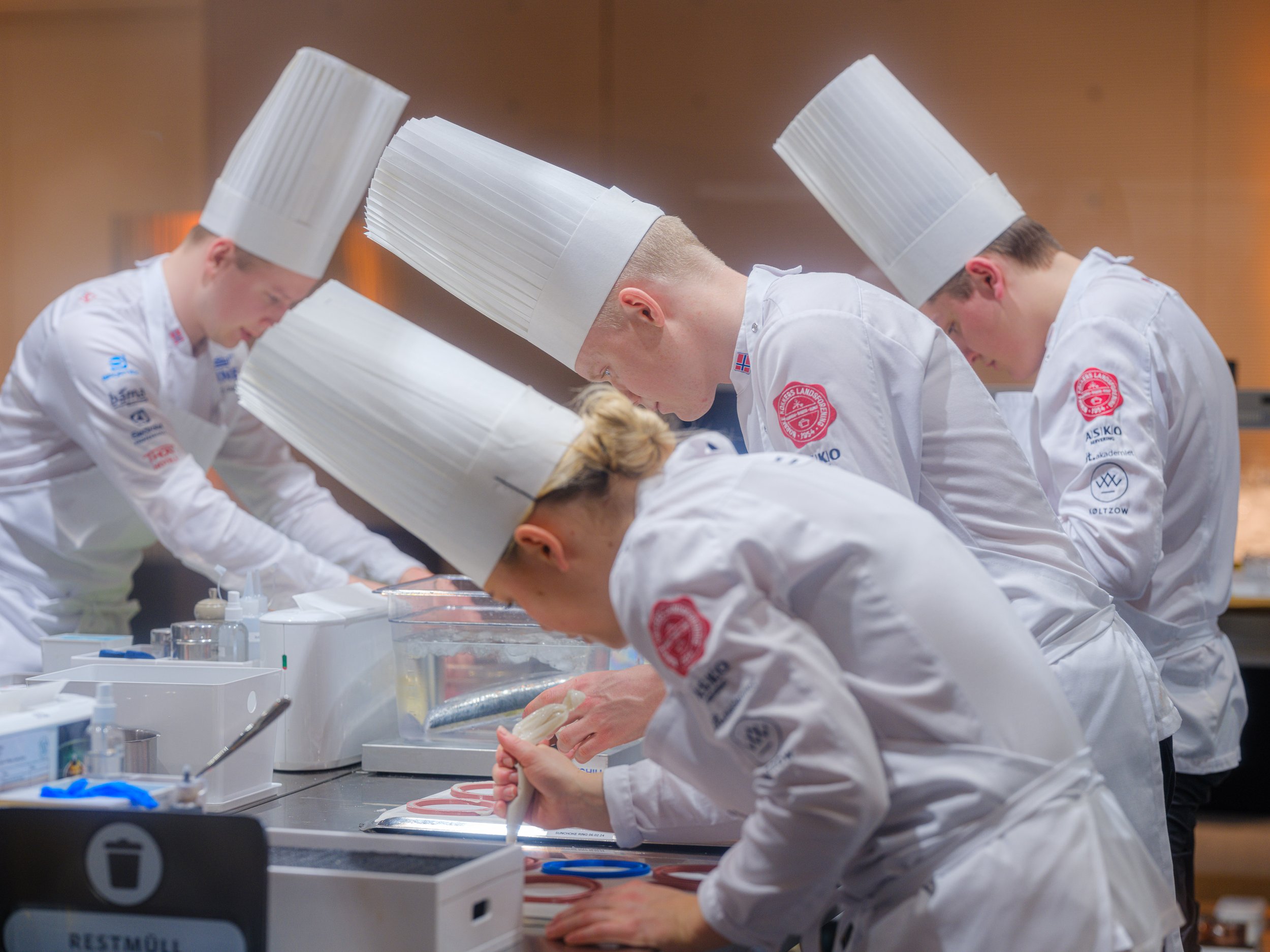 Fem kokker i hvite uniformer og høye kokkehatter som jobber i et kjøkken.