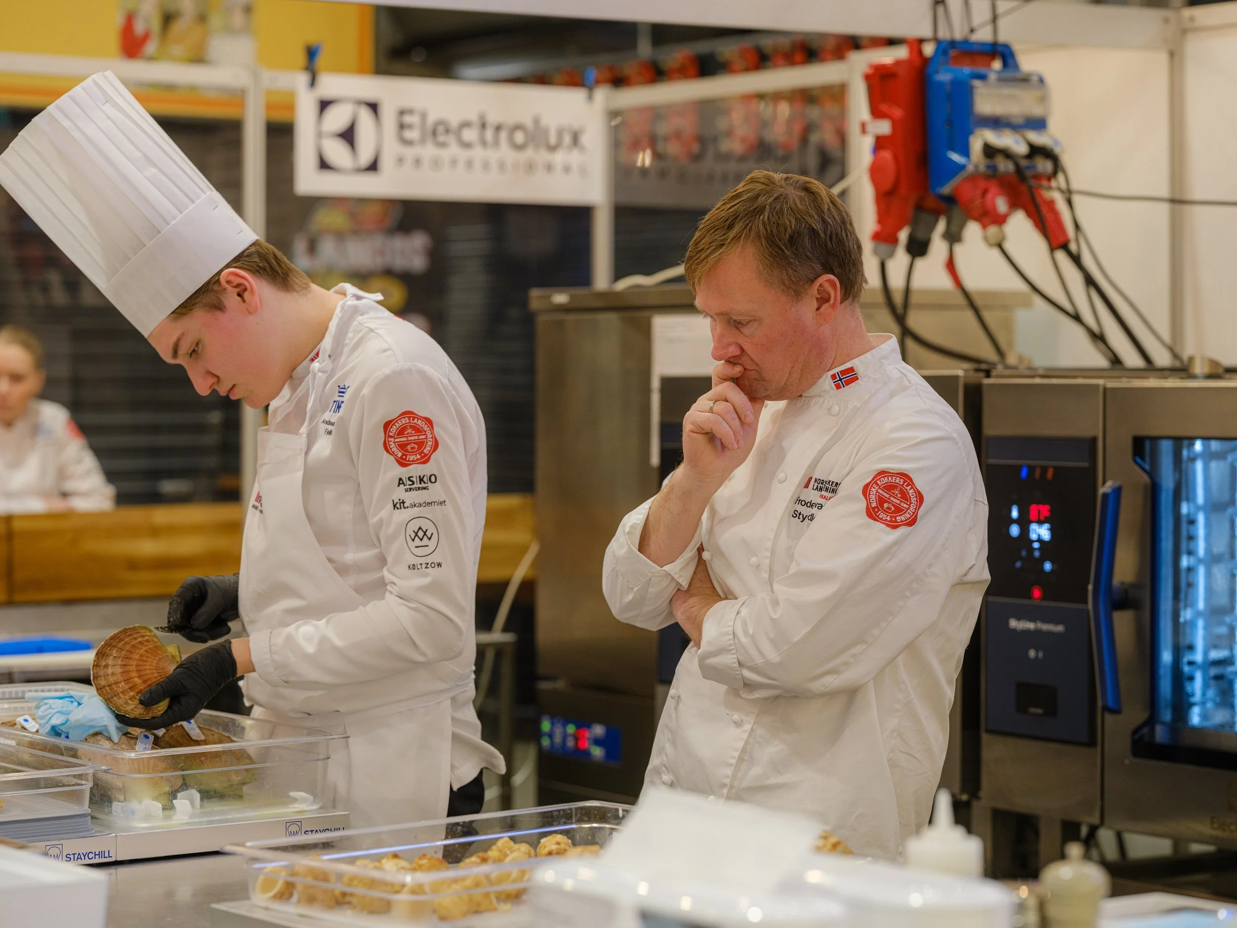 To kokker, en yngre kvinne og en eldre mann, jobber i et kjøkkenmiljø. Den yngre kvinnen har på seg en kokkehatt og jobber med mat, mens den eldre mannen ser ut til å vurdere noe nøye.