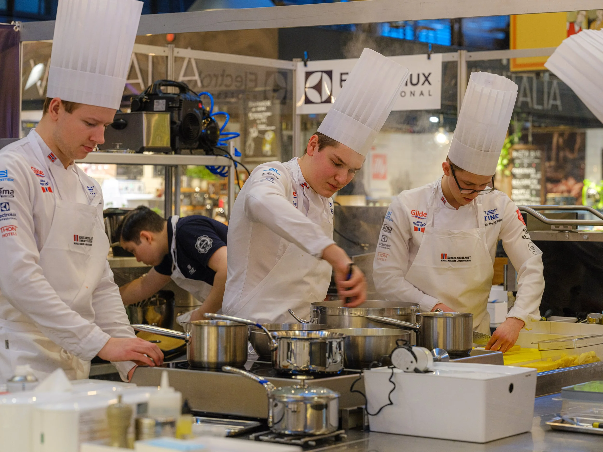 Fire kokker i hvite uniformer og kokkehatter lager mat i et kjøkken, med metallkasseroller og kjøkkenutstyr.