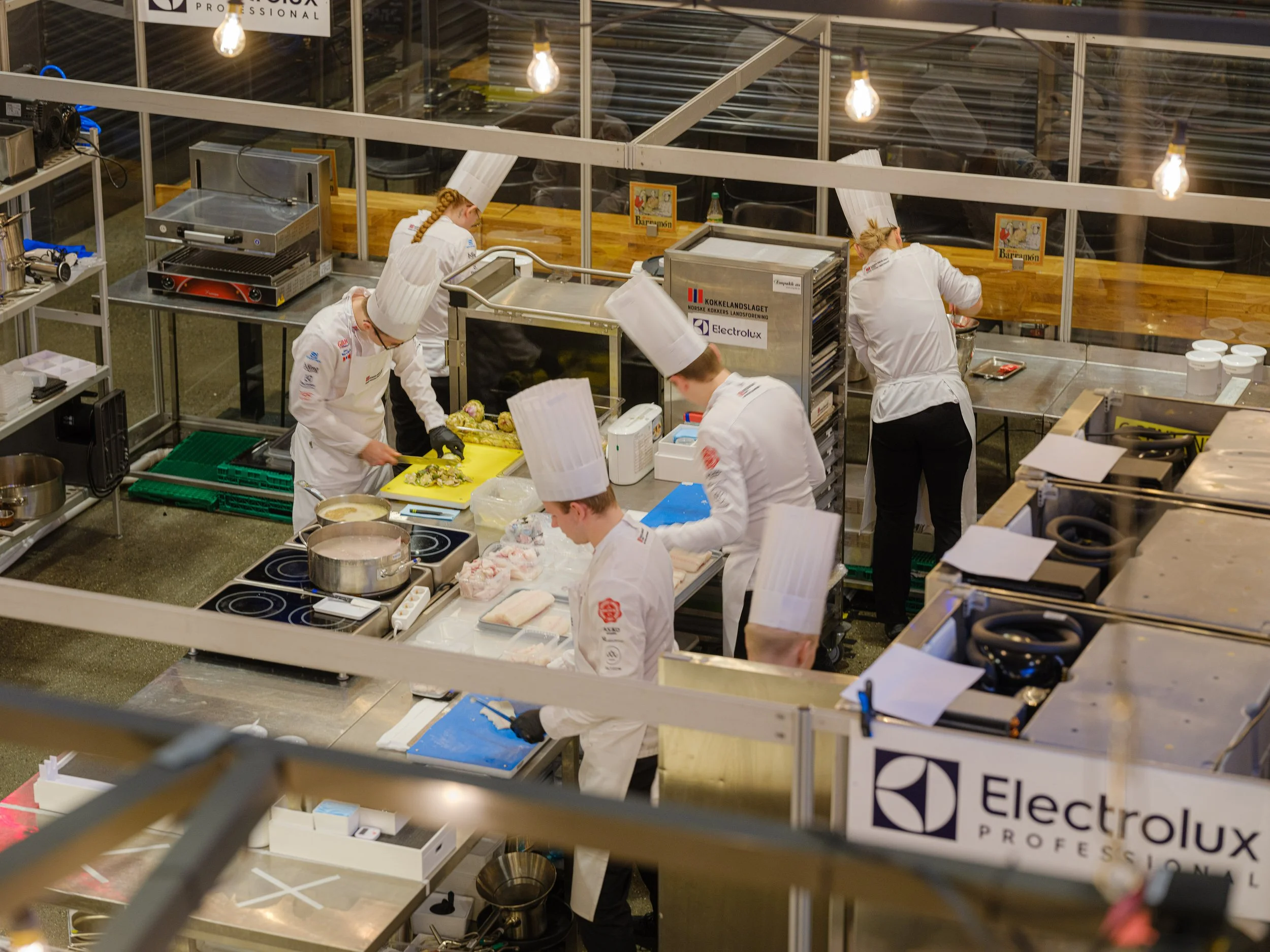 Kokker som forbereder mat i et profesjonelt kjøkken, med kokeutstyr, skjærebrett og andre kjøkkenredskaper, med Electrolux branding i bakgrunnen.