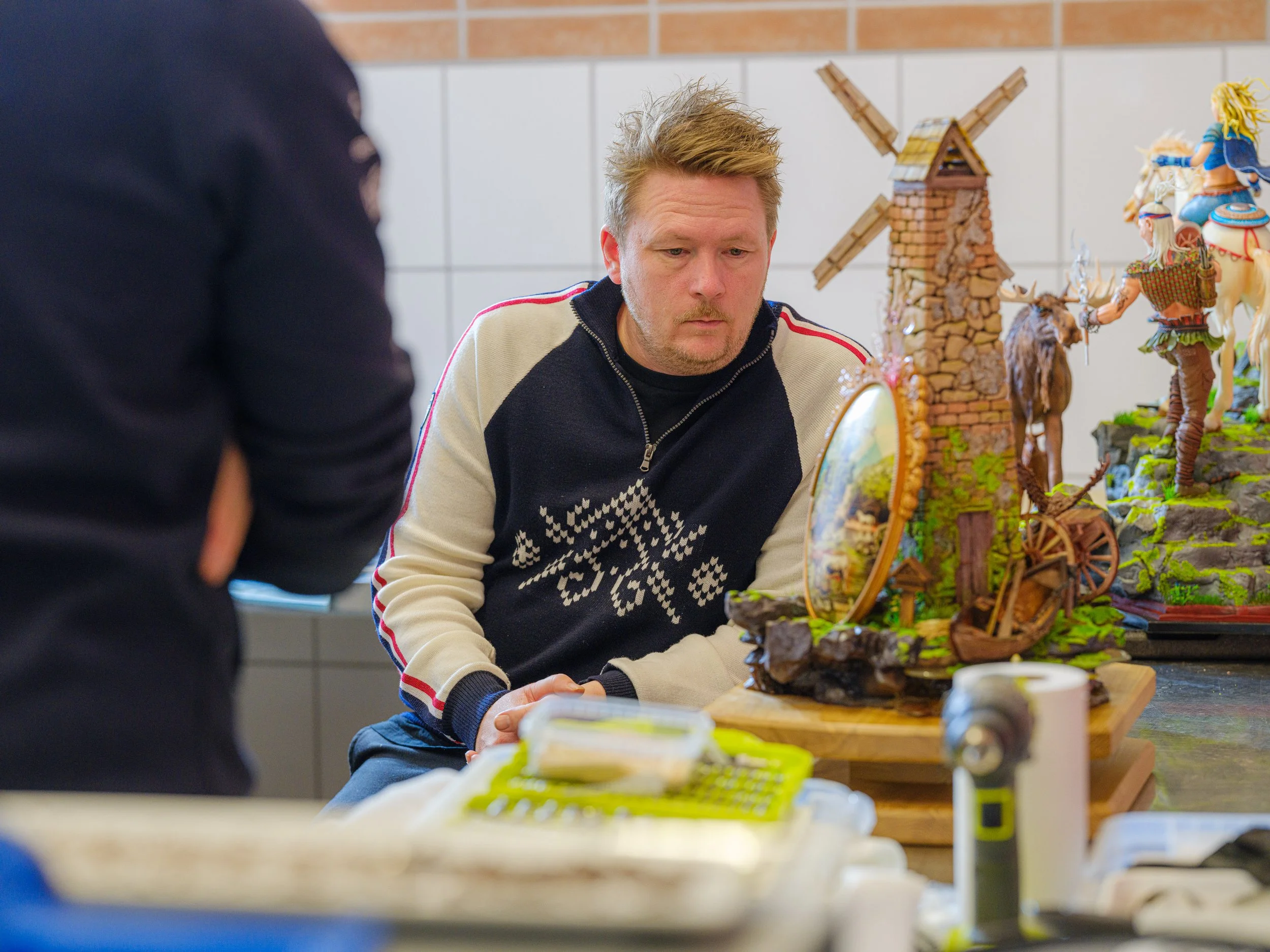 En mann ser på en detaljert modell av et landskap med vindmølle, hus, en hest og figurer, plassert på et bord.