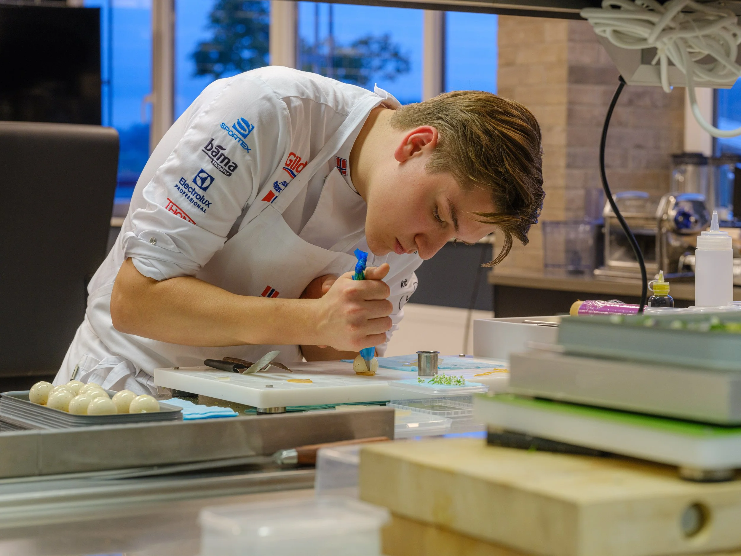 En ung mann med hvit kokkejakke pyntet med norske flagg, som jobber med dekorering av en kake i et kjøkken.