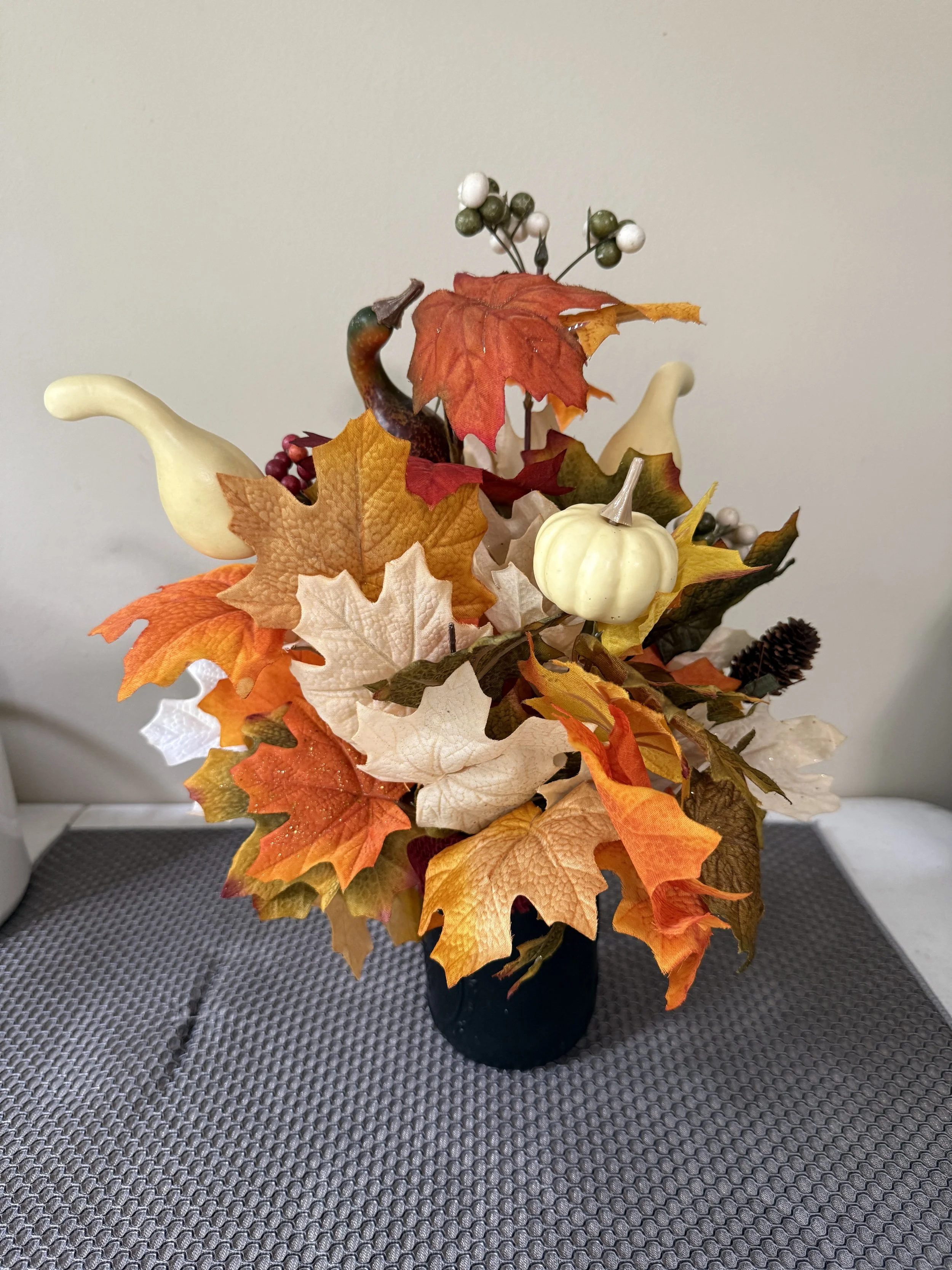 Fall-themed artificial floral arrangement in a black vase, featuring white pumpkins, white and green berries, and colorful autumn leaves in shades of orange, red, yellow, and beige.