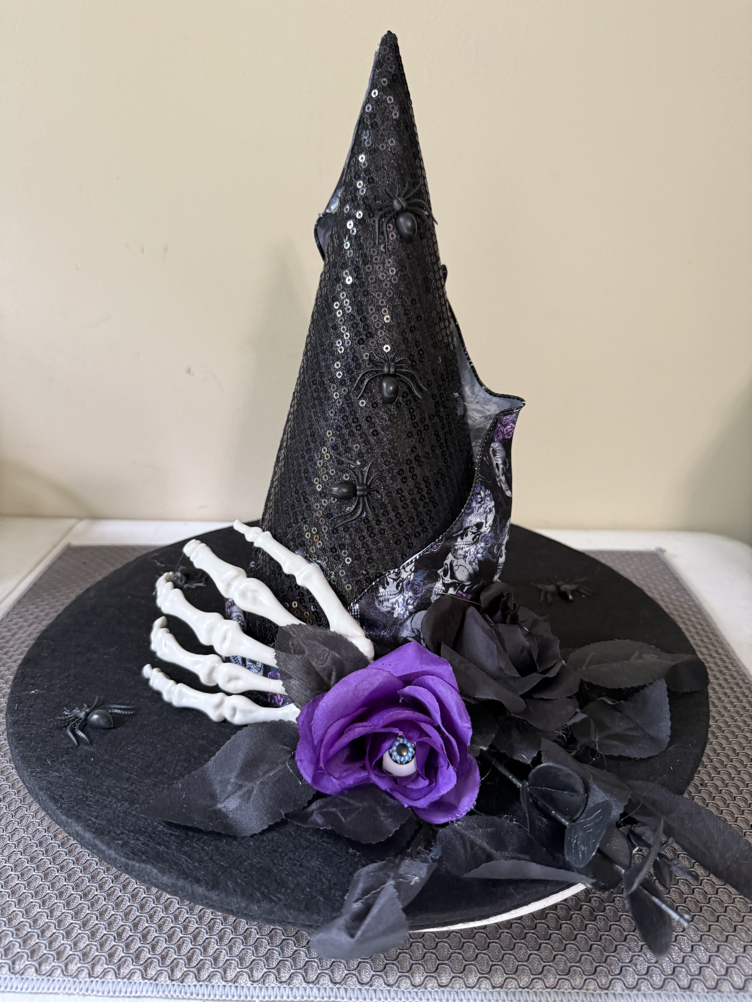 A Halloween-themed cake decorated with a black witch hat, black spiders, a purple rose with an eyeball in the center, black leaves, and black insects, all on a black circular base.