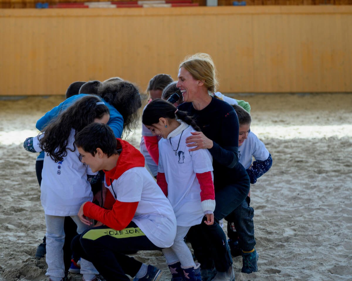 Smiling instructor hugging a group of laughing children gathered around her on the sand arena of Ivó Riding Hall during a group activity at a children's riding program.