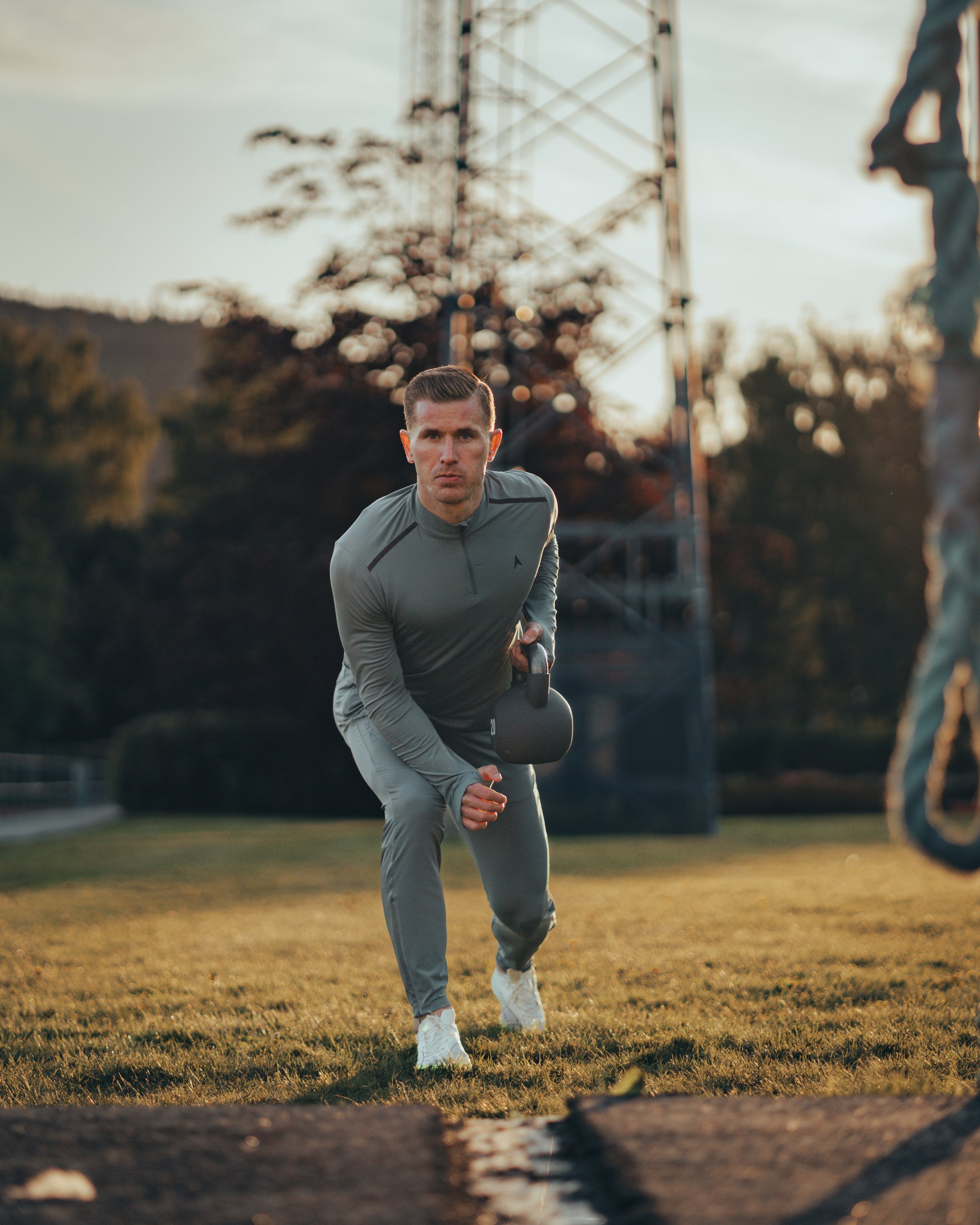 En mann som trener utendørs med en kettlebell på en gressplen. Han bærer treningstøy og står midt i en øvelse. Bakgrunnen viser noen trær og en struktur med stålkonstruksjon.