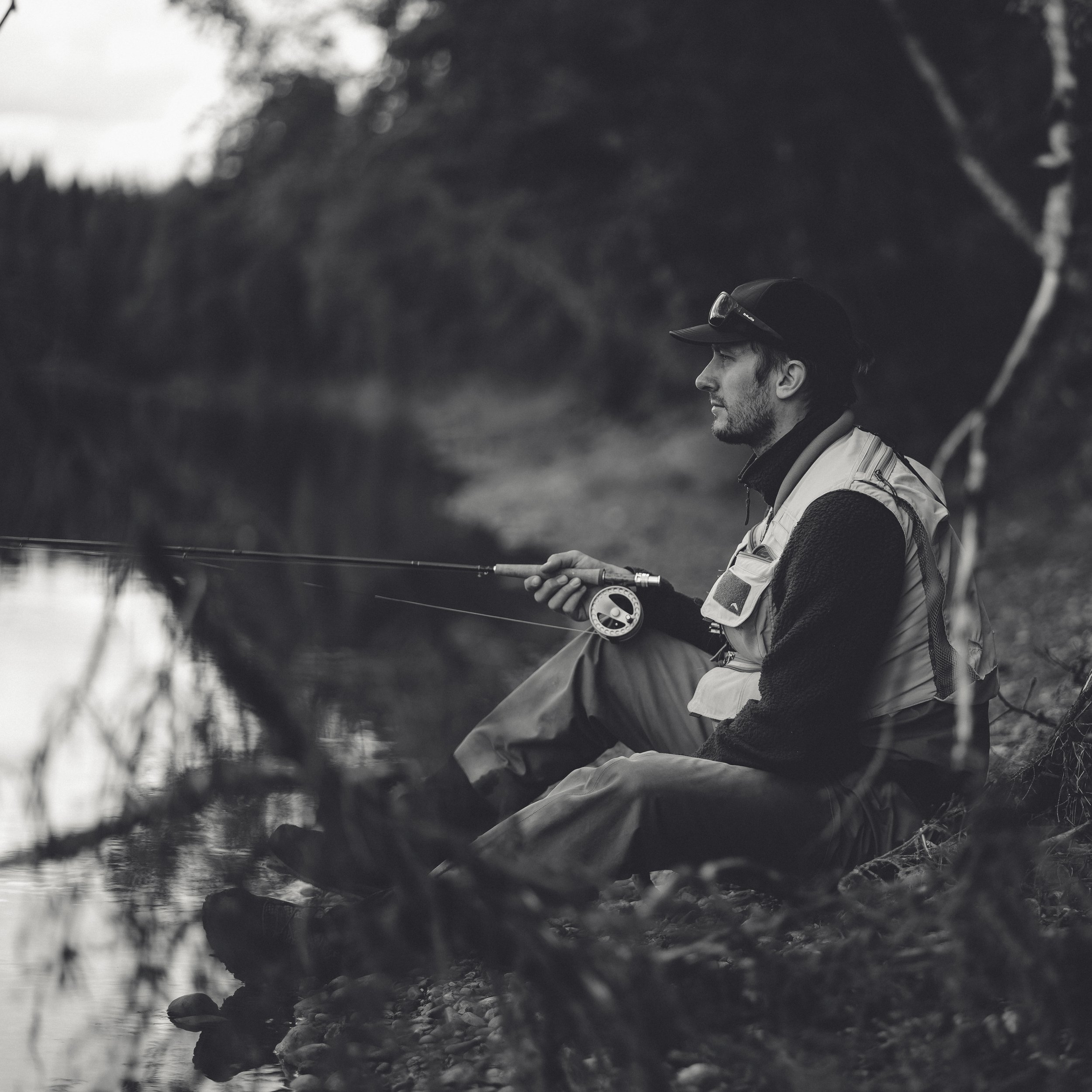 En mann sitter ved en elv og fisker med en fiskestang, iført en fiskervest og caps, omgitt av natur.