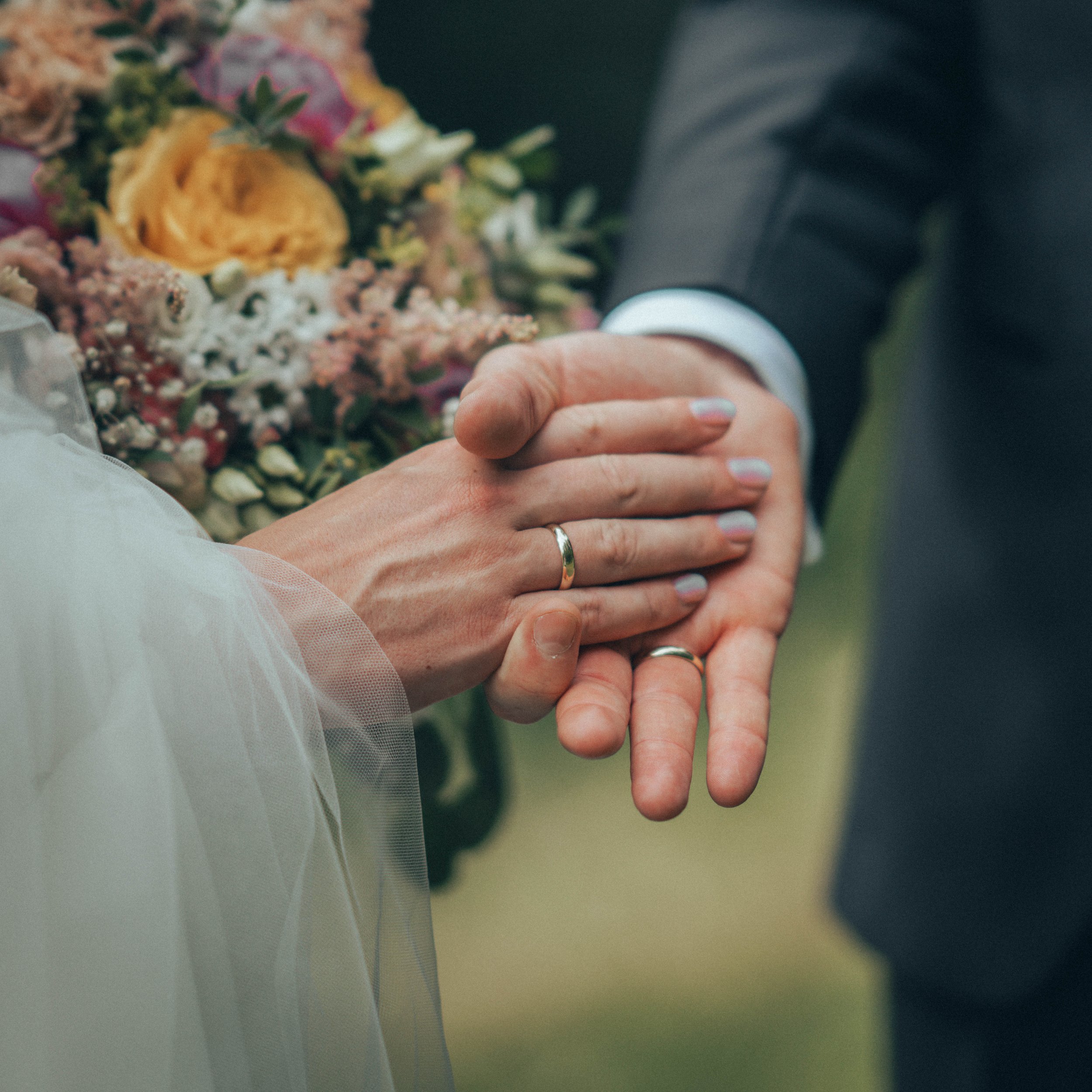 Brudepar som holder hender med bryllupsringer, blomsterbukett i bakgrunnen.