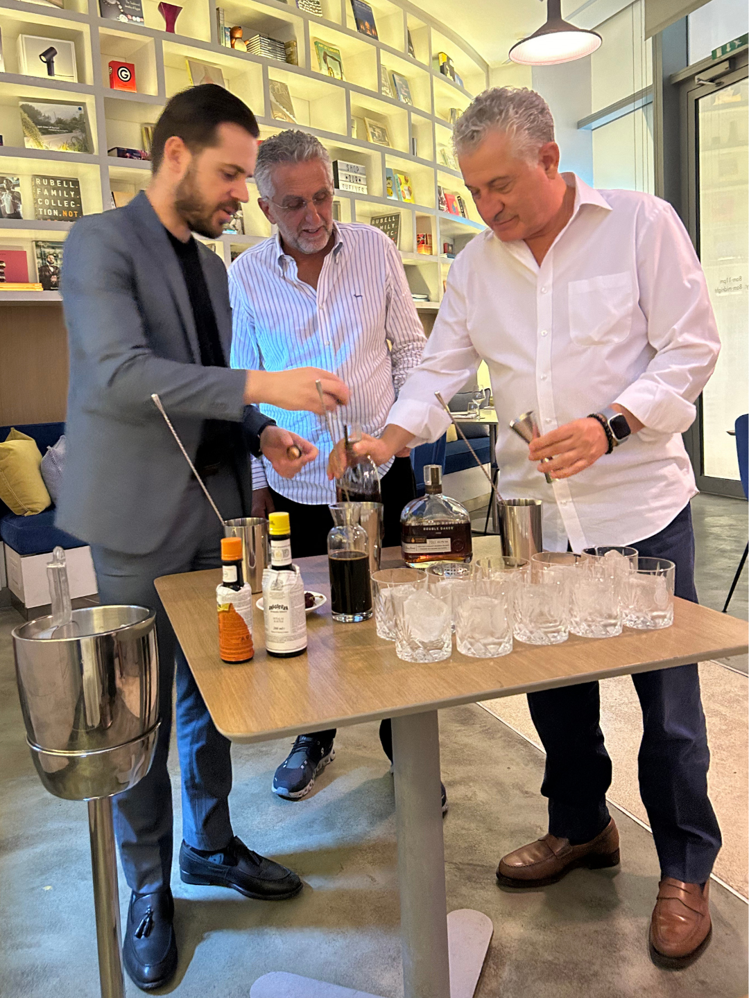 Three men are preparing whiskey at a table with various bottles and glasses in a modern indoor setting.