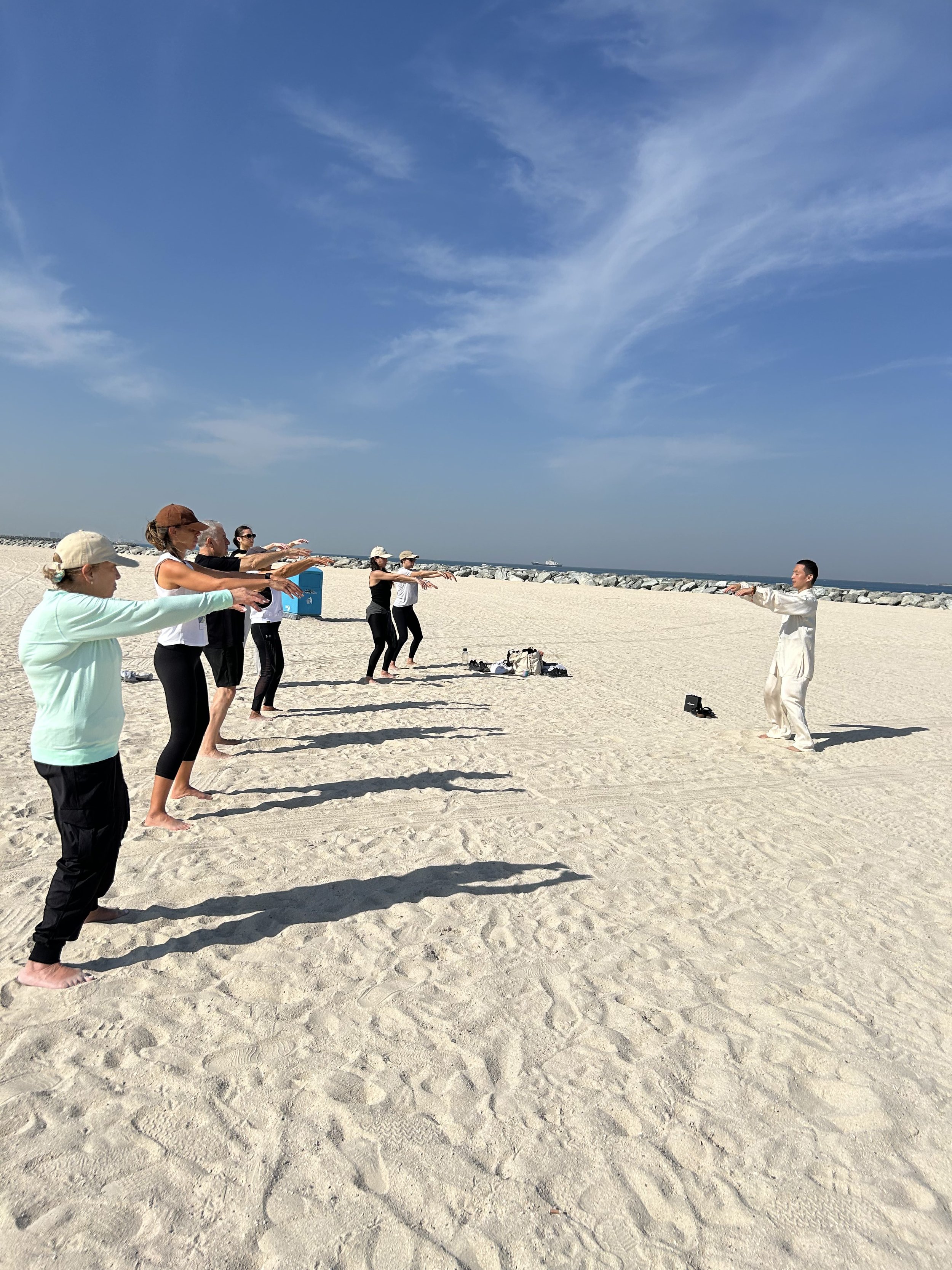 Group of people practicing Tai Chi on a sandy beach under a clear blue sky.