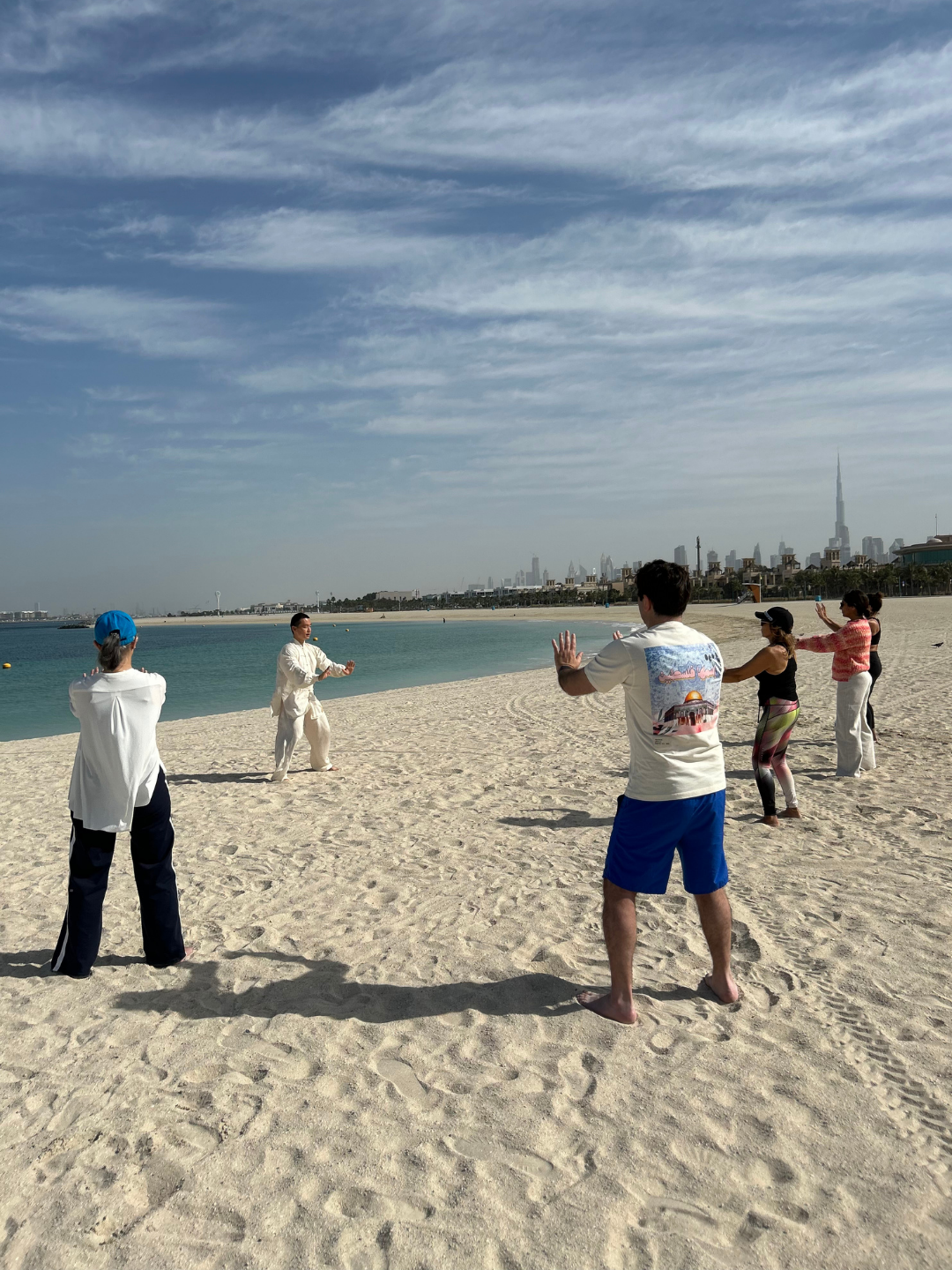 Group of The Nadi members practicing Tai Chi a sandy beach with the Dubai city skyline in the background and cloudy sky overhead.