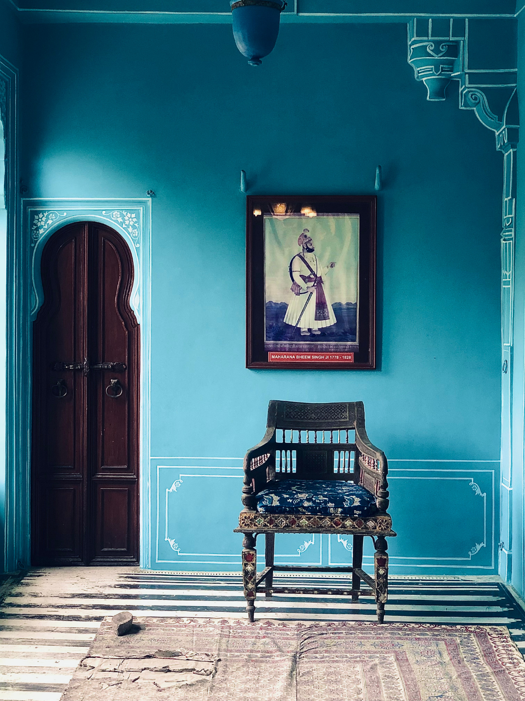 A room with a blue wall featuring white decorative trim. There is a wooden door with an arched top on the left and a framed portrait hanging on the wall. A wooden chair with a patterned cushion sits in front of the wall, and a small rug with a torn section is on the floor.