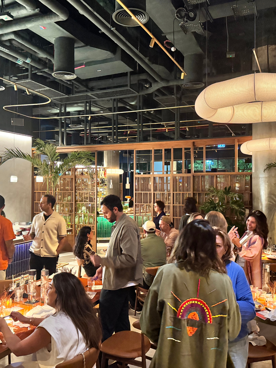 People dining and socializing at a The Nadi event, in a modern restaurant with a dark ceiling, hanging lights, and wooden partitions, along with potted plants and ambient lighting.
