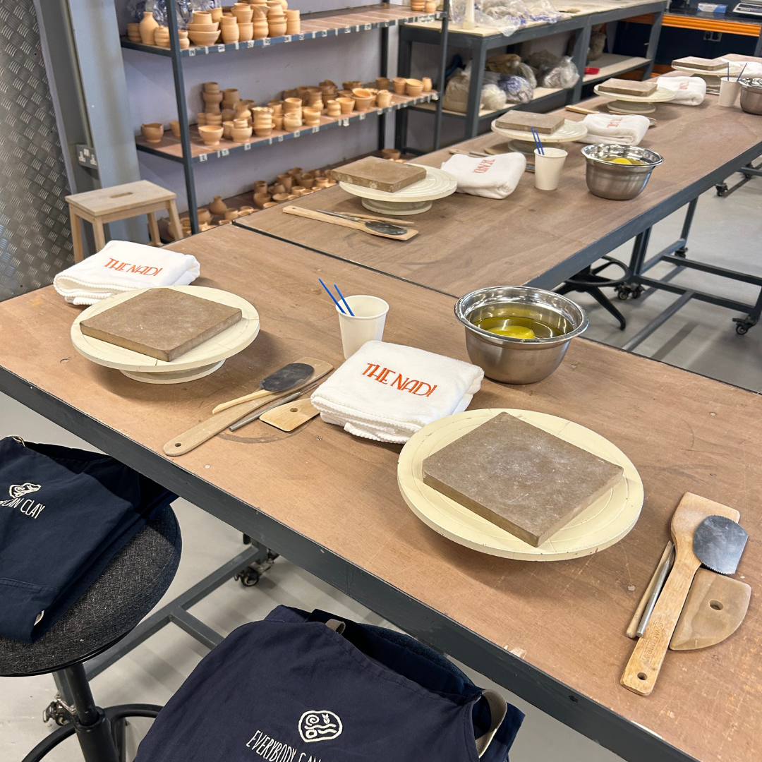 A pottery class setup with pottery tools and 'The Nadi' branded merchandise. Shelves in the background hold numerous unglazed clay pottery pieces.