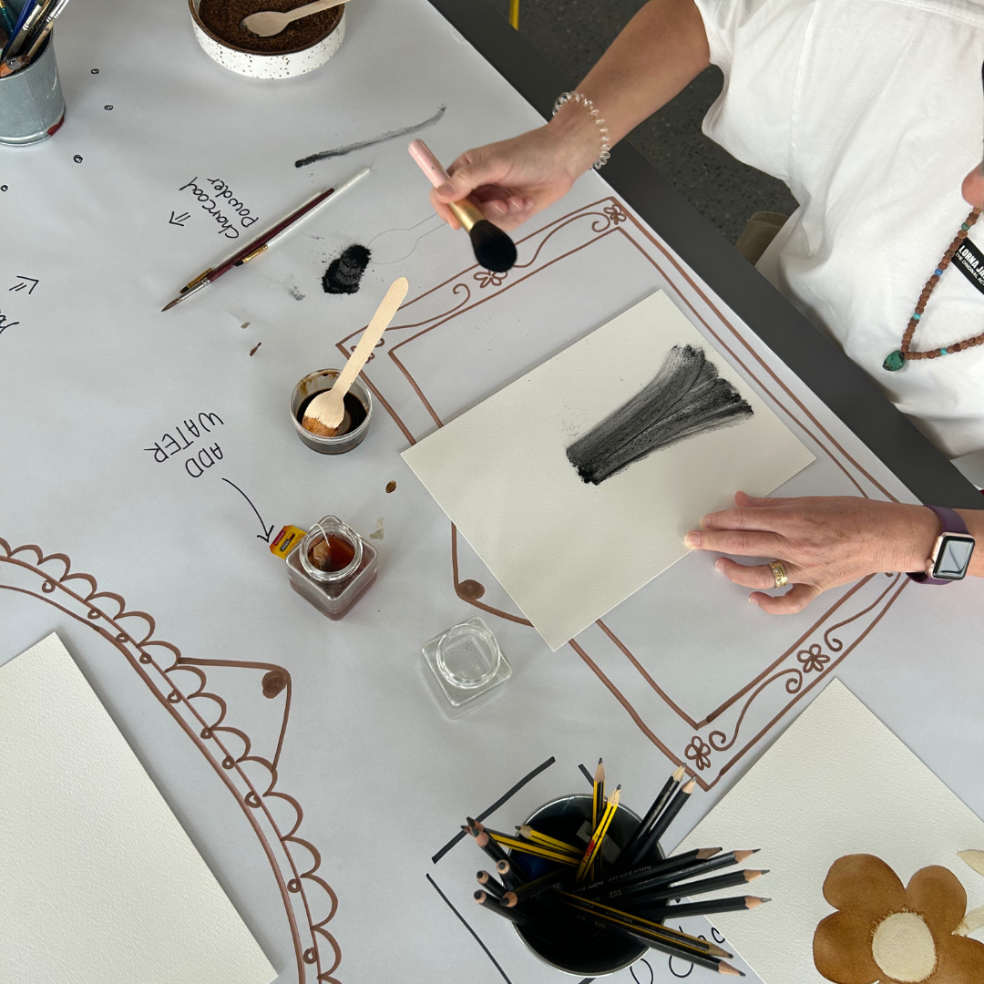 Person holding a makeup brush over a sheet of paper with black paint strokes, surrounded by coffee and tea supplies, pencils, and art notes on a table.