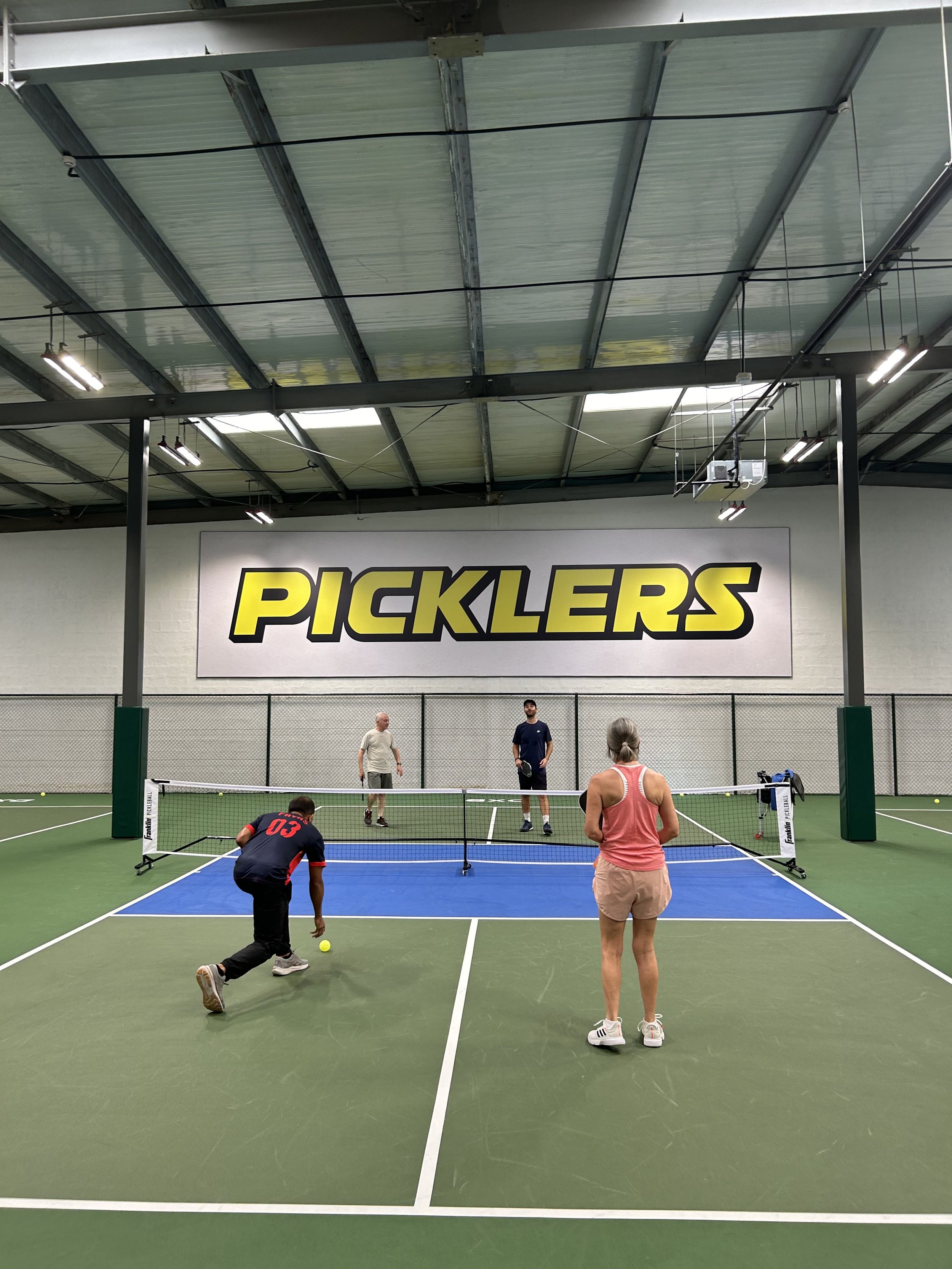 Indoor pickleball game with four players on a court, featuring a large "Picklers" sign on the wall in the background.