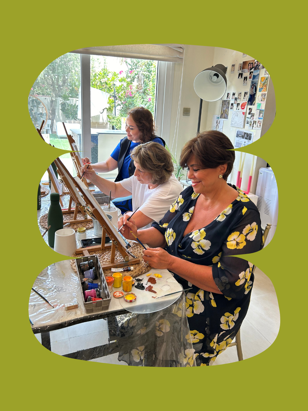 Three women painting on canvases set on easels at a table in a well-lit room with a large window showing a garden outside. The women are focused and smiling and are at a painting event organised by The Nadi.