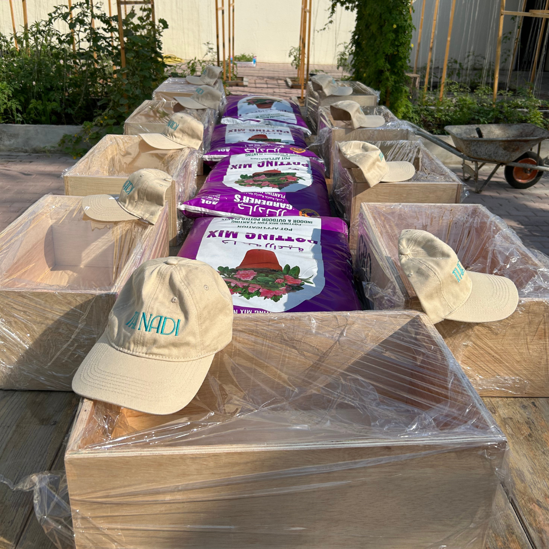 Picture of a gardening event, showing garden boxes with gardening compost along with beige caps with 'The Nadi' embroidered on them, outdoors on a wooden table covered with clear plastic, garden in background, wheelbarrow nearby.