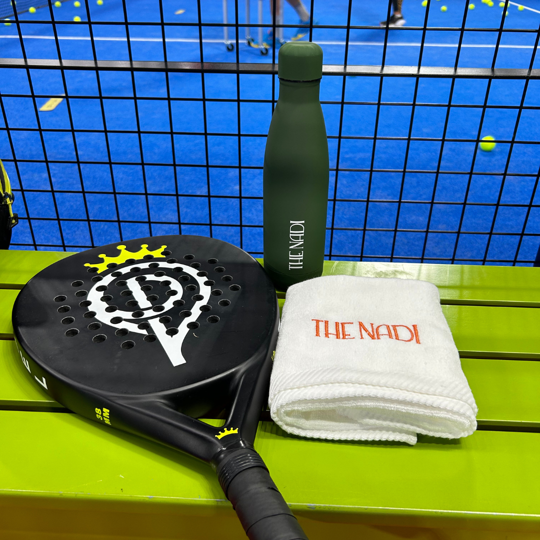 Padel tennis racket, green water bottle, and white towel with 'THE NADI' embroidered in orange, placed on a green bench at an indoor padel tennis court.