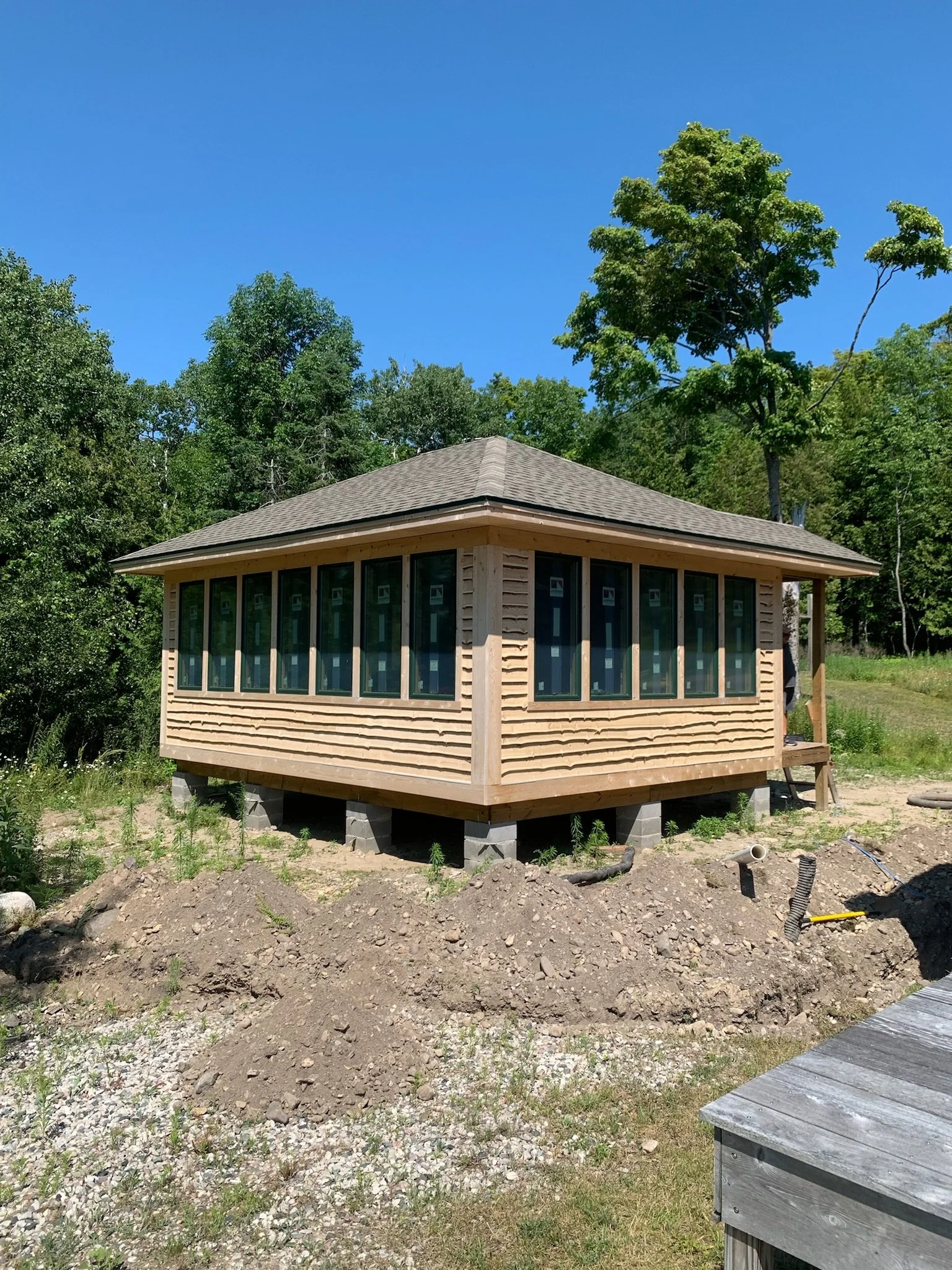 A small wooden house or cabin under construction with a raised foundation and a shingled roof, surrounded by trees and dirt, on a clear sunny day.