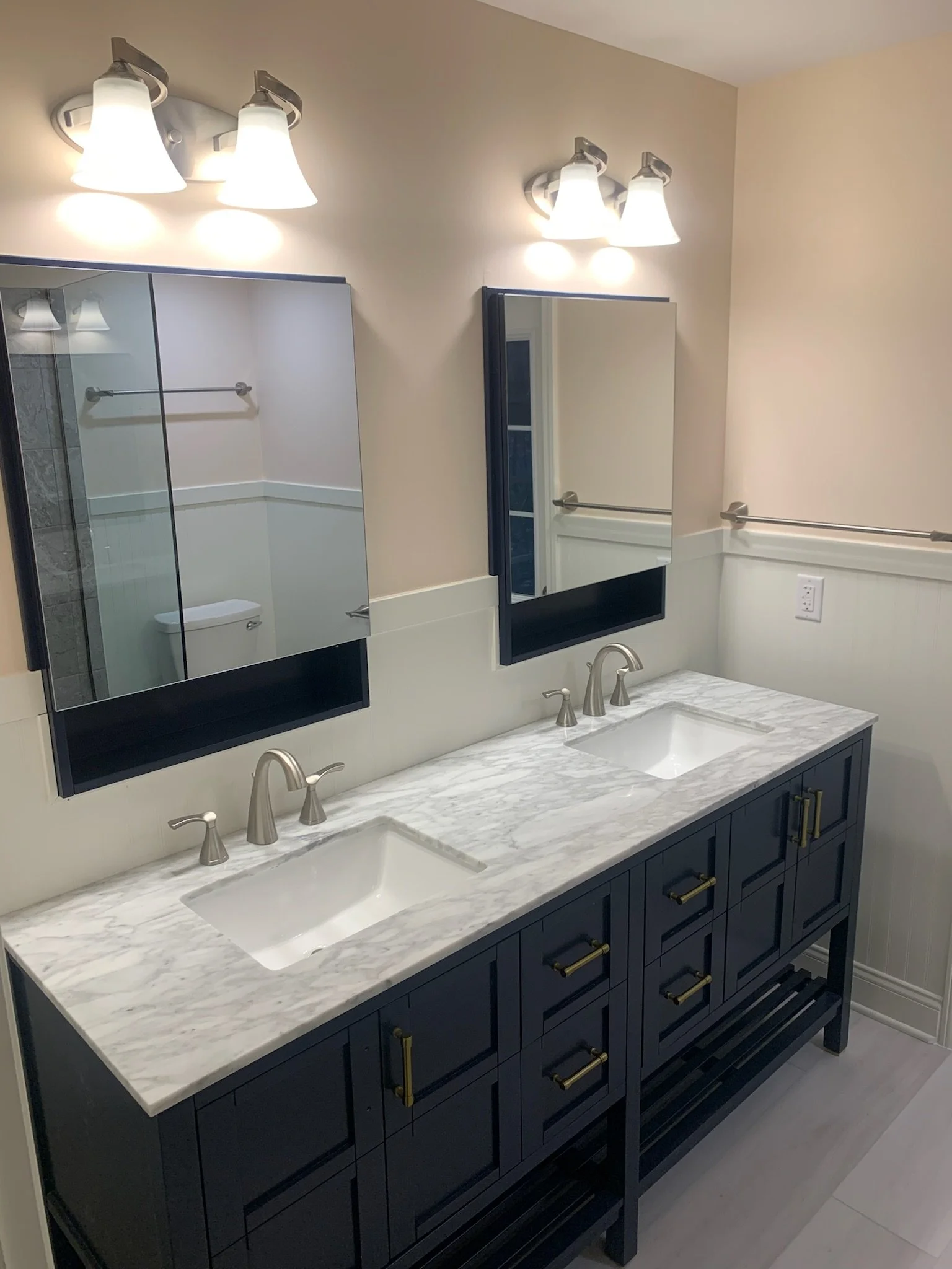 Bathroom with double vanity featuring a white marble countertop, dark navy cabinets with gold handles, two mirrors, and three-light fixtures above each mirror.