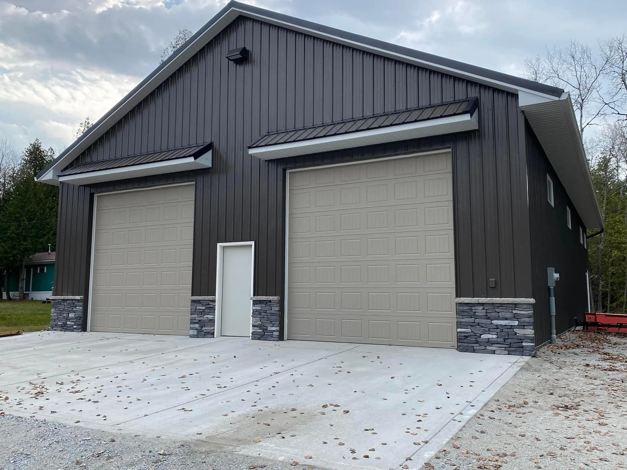 A modern black metal building with two large beige garage doors, a small side door, and a stone foundation, situated on a concrete driveway with scattered leaves.