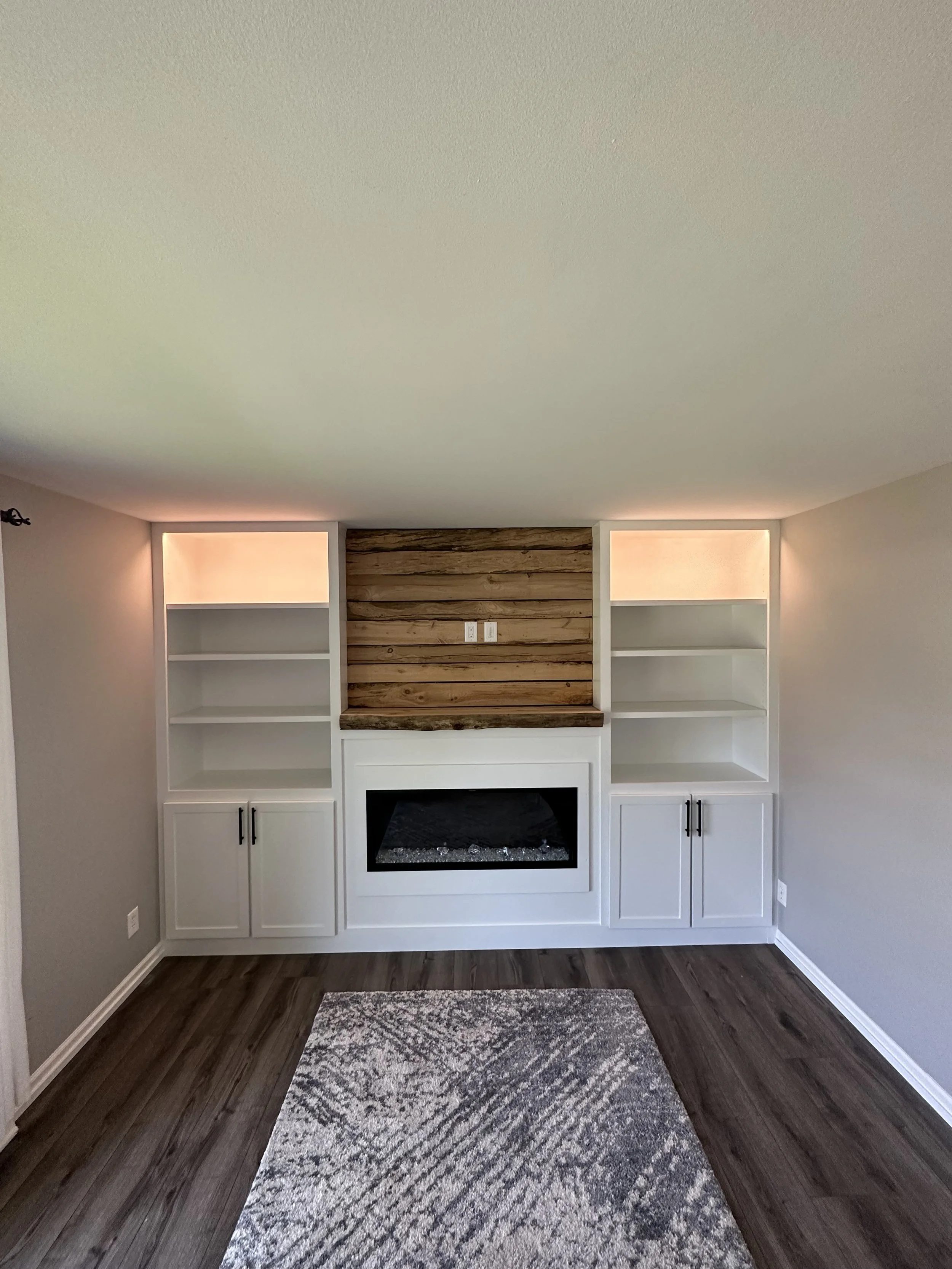 Living room with built-in white bookshelves on each side, a wood-paneled accent wall, electric fireplace and a patterned rug on dark hardwood floor.