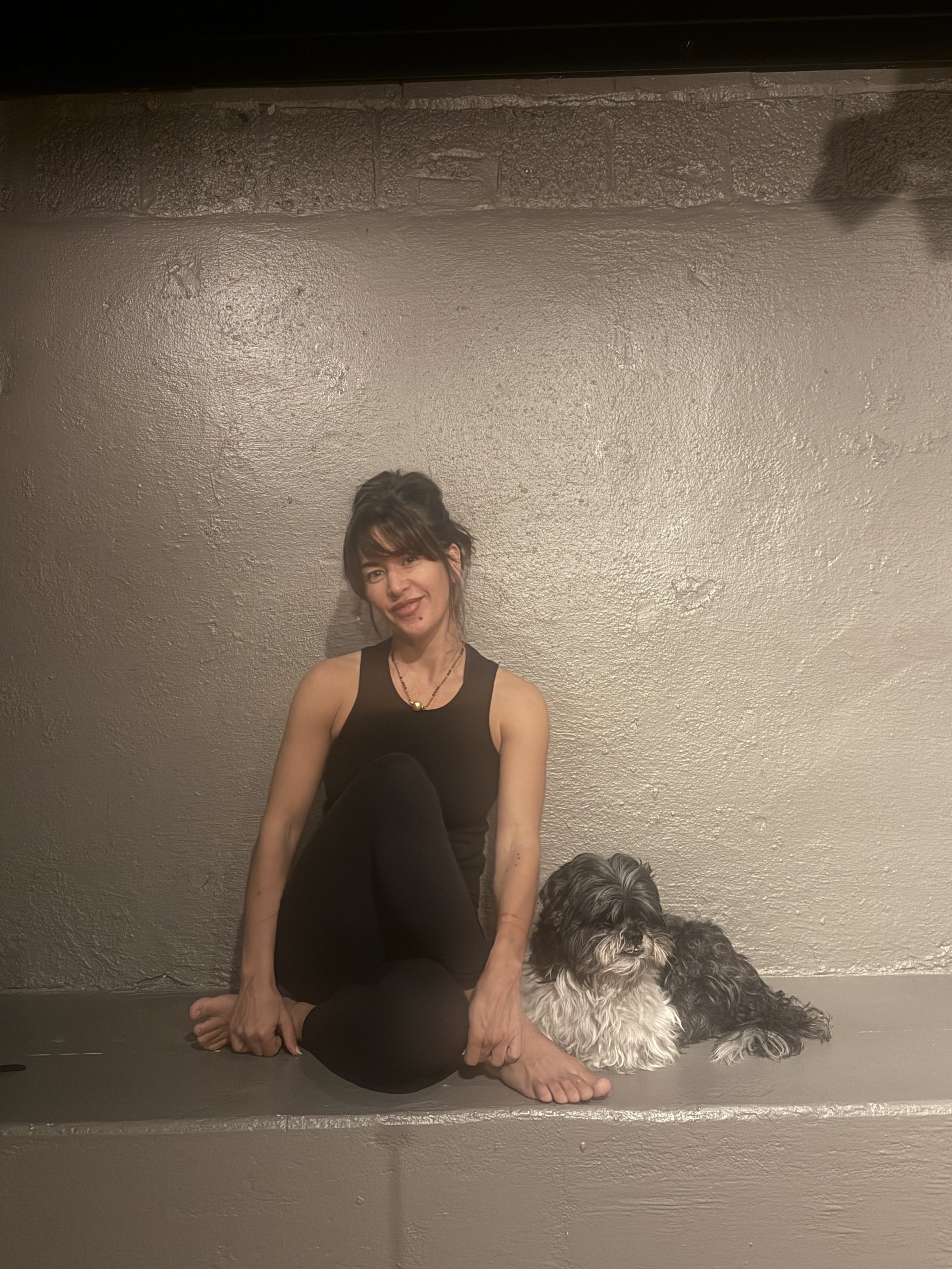 A woman practicing yoga with a dog sitting in front of her, both on a gray floor against a gray wall.