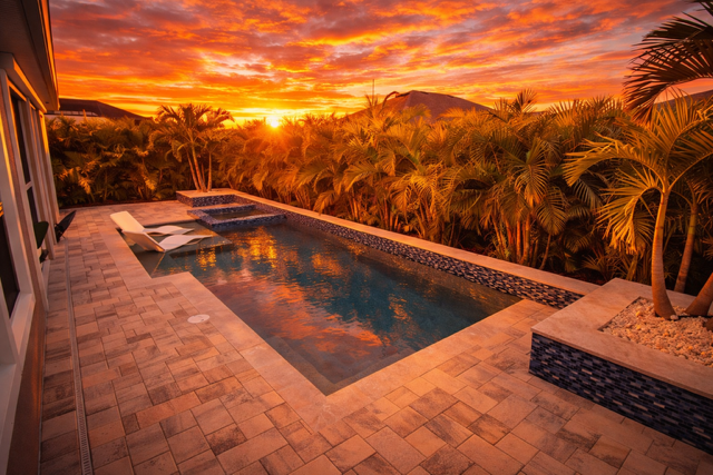 A backyard pool during sunset, surrounded by palm trees and a paved patio.