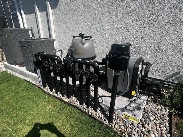 Outdoor solar-powered pool or spa filtration system with pumps and electrical components installed along a house exterior wall, surrounded by rocks and grass.