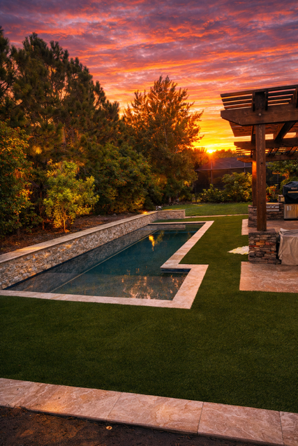 A backyard swimming pool at sunset with surrounding trees, a grassy area, and a wooden patio structure with outdoor furniture.