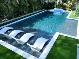 A modern backyard swimming pool with four white lounge chairs on a grassy area, surrounded by lush green plants.