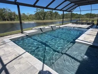 A screened outdoor pool area with lounge chairs along the poolside, overlooking a body of water and green trees in the background.
