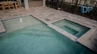 A swimming pool and a hot tub in a backyard with a tiled deck and a fence.