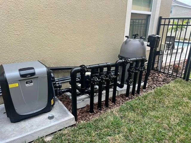 Pool pump system with black pipes, a large grey filtration tank, and a control box, installed next to a grass lawn and a beige house wall.