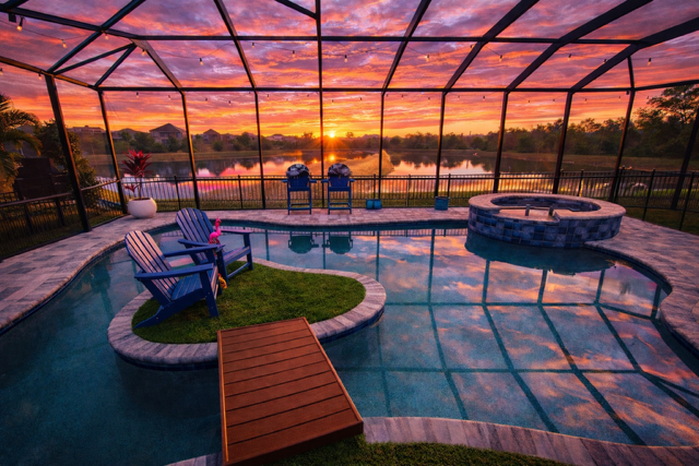 Enclosed backyard pool area with two blue Adirondack chairs near the pool, a hot tub, and a sunset over a lake in the background.