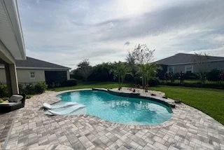 Backyard with a kidney-shaped swimming pool, poolside lounge chairs, well-maintained grass, and houses in the background under a partly cloudy sky.