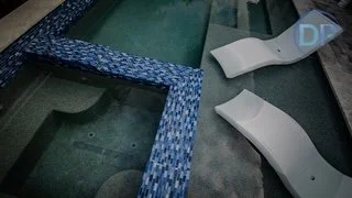 Corner of a swimming pool with a blue mosaic tile border and empty white lounge chairs.