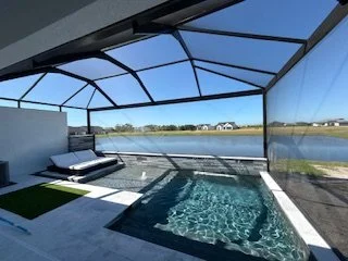 Indoor pool area with a hot tub, enclosed by glass walls and ceiling, overlooking a lake and grassy landscape.
