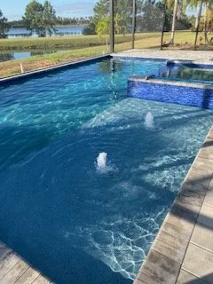 Outdoor swimming pool with jets creating water bubbles, surrounded by a paved deck and a scenic view of trees and a body of water in the background.