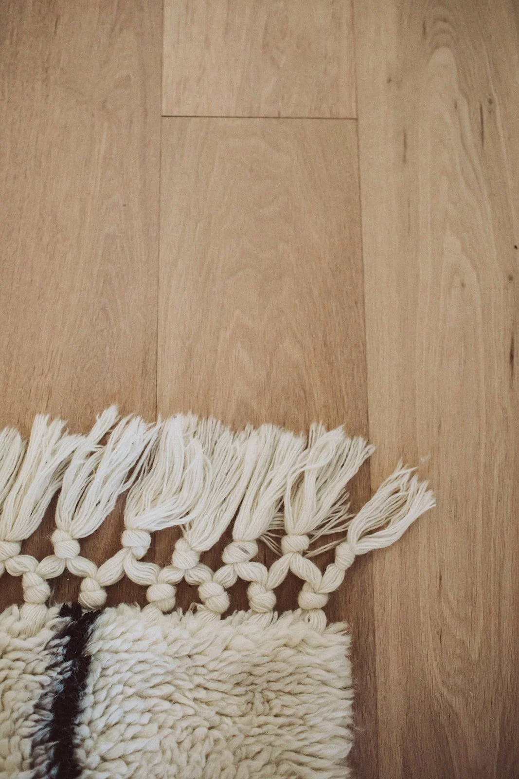 Close-up view of a light-colored wooden floor and a corner of a beige and black fluffy rug with tassels.